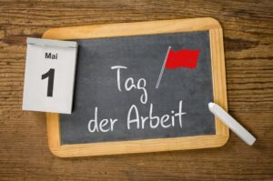 Chalkboard displaying the German phrase 'Tag der Arbeit' with a small red flag illustration, on a wooden table; a May 1 calendar is clipped to the side.