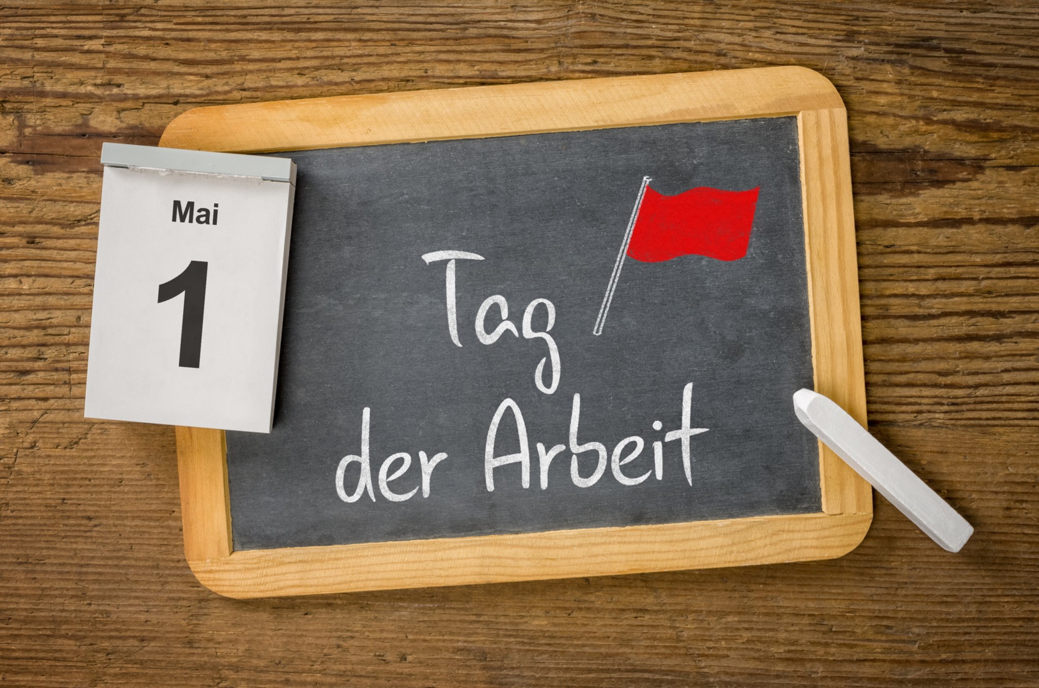 Chalkboard displaying the German phrase 'Tag der Arbeit' with a small red flag illustration, on a wooden table; a May 1 calendar is clipped to the side.